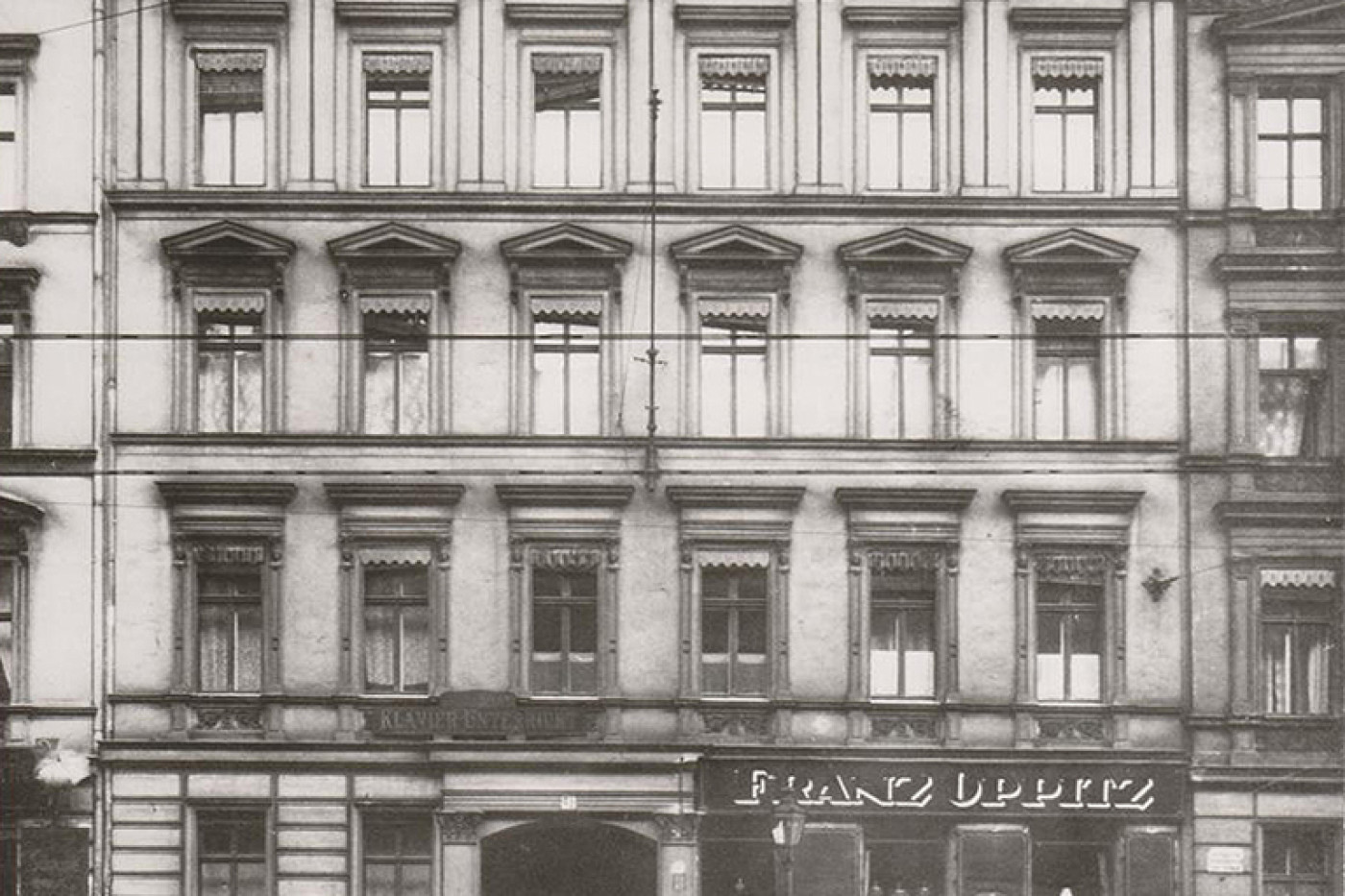 Historic black and white photograph of the building with its stucco fa&ccedil;ade, round-arched portal and a shop on the ground floor.