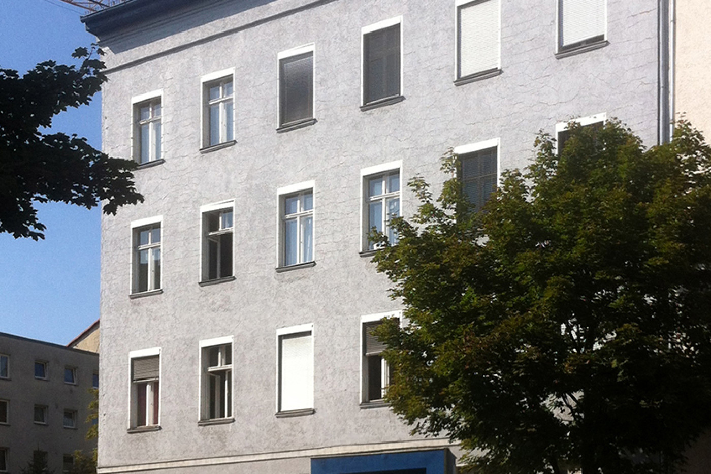 The building before the roof extension: plain grey render with a blue-painted ground floor, parked cars in front.