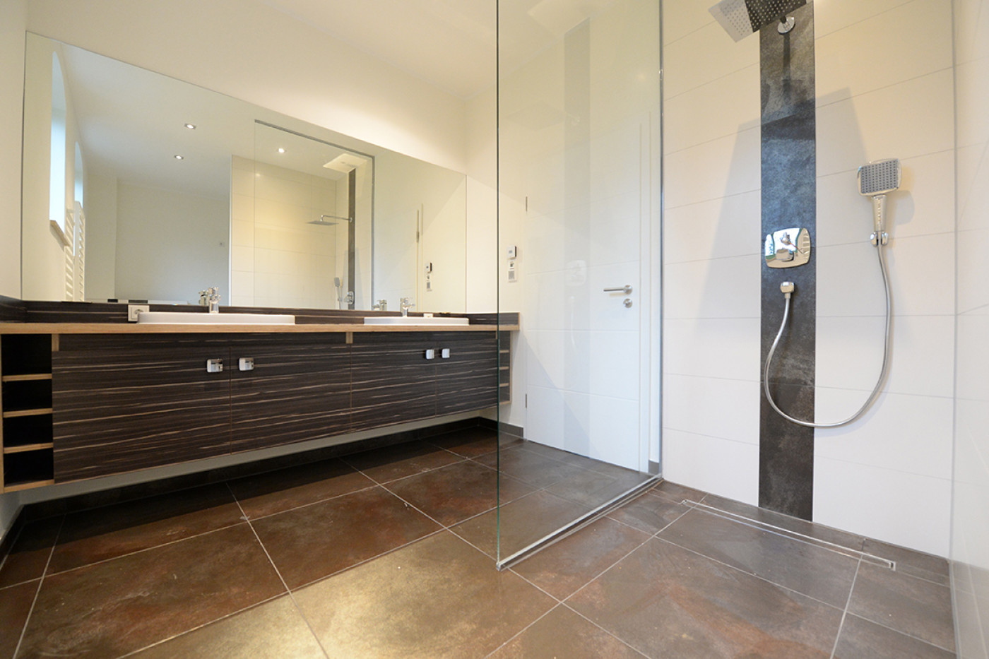 Bad mit bodengleicher Dusche hinter Glaswand, Doppelwaschtisch auf dunklem Unterschrank und gro&szlig;formatigen Bodenfliesen.
