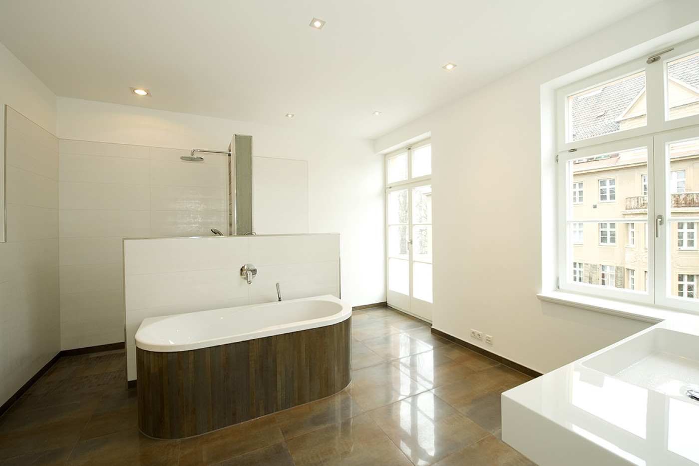 Helles Bad mit dunklem Steinboden, holzverkleideter Wanne und Dusche, daneben hohe Fenster zum Hof.