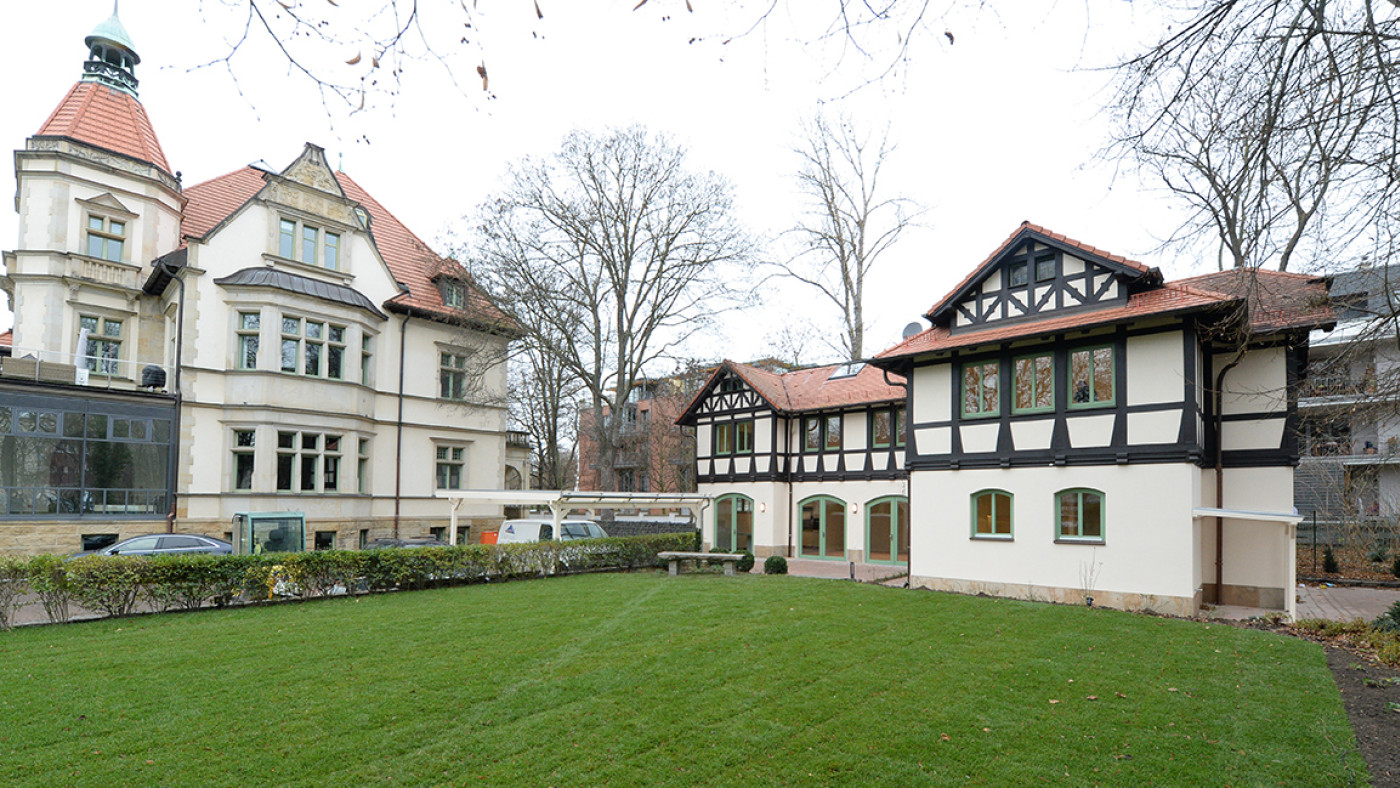 Denkmalgesch&uuml;tzte Villa mit Turm, rechts daneben das Kutscherhaus mit Fachwerkgiebel, davor eine Rasenfl&auml;che.