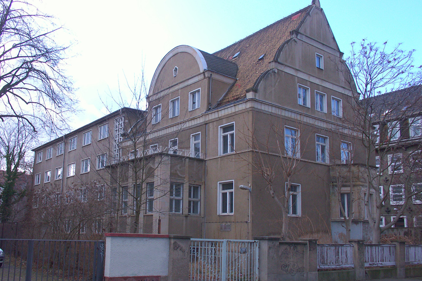 Die Villa vor der Sanierung: verwitterter Putz, teils zugesetzte Fenster, davor ein Zaun und kahle B&auml;ume.
