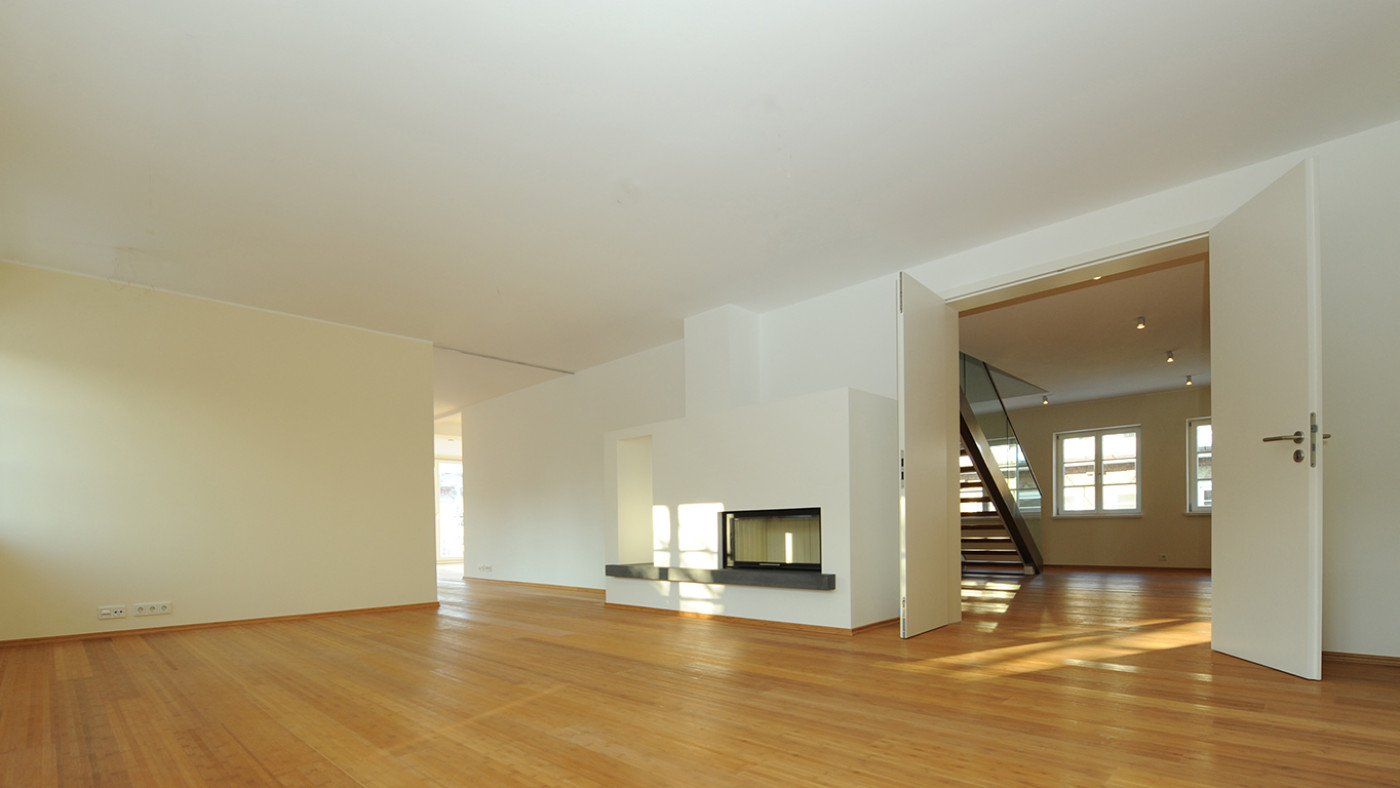 Leerer sanierter Wohnraum mit Parkettboden, zweiseitigem Kamin und Blick zum Treppenhaus.