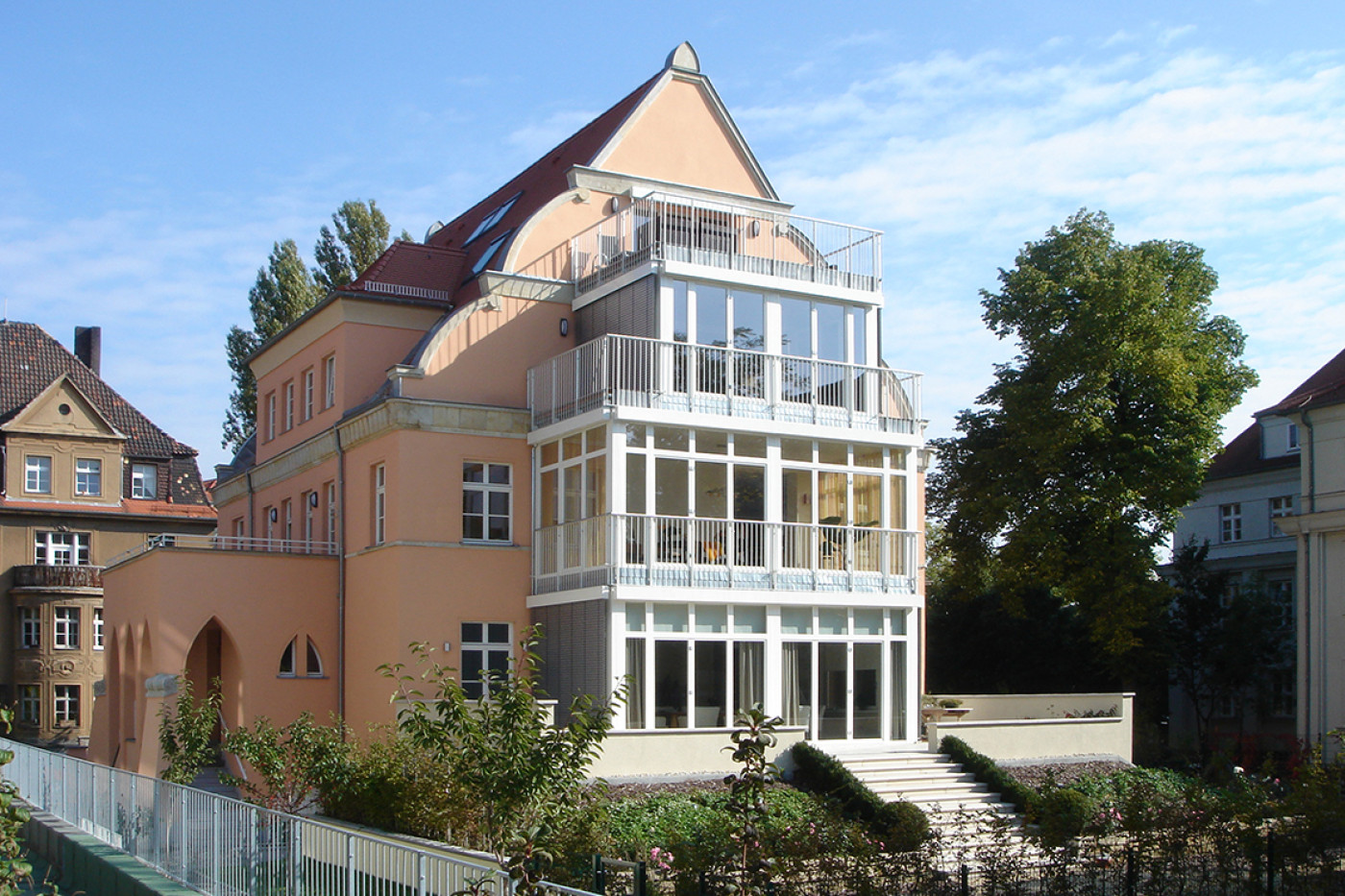 Die sanierte Villa mit lachsfarbenem Putz, geschwungenem Giebel und wei&szlig;em verglastem Anbau &uuml;ber drei Geschosse.
