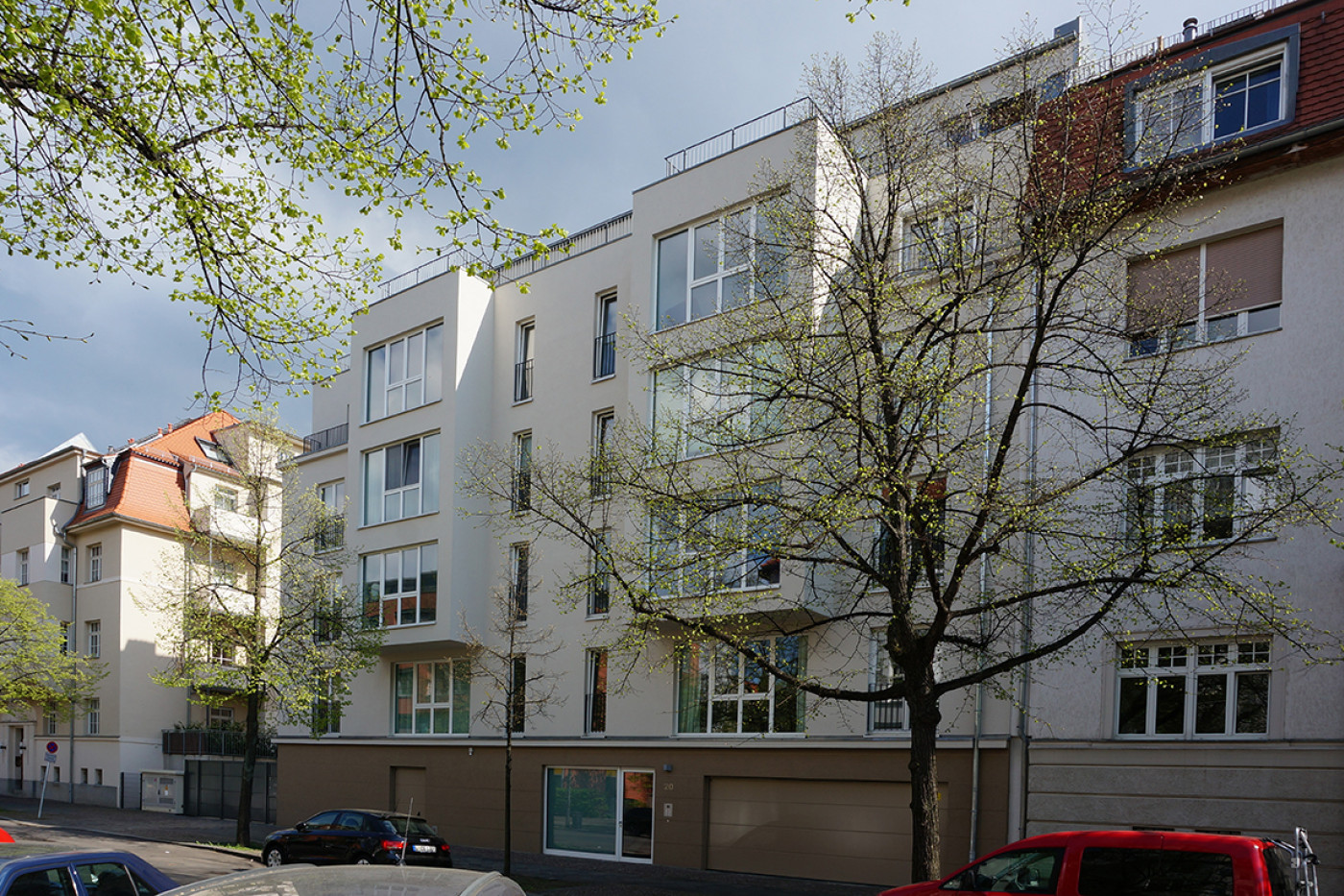 Neubauteil mit hellem Putz, Balkonen und Dachterrasse zwischen Altbauten, davor eine Stra&szlig;e mit parkenden Autos.