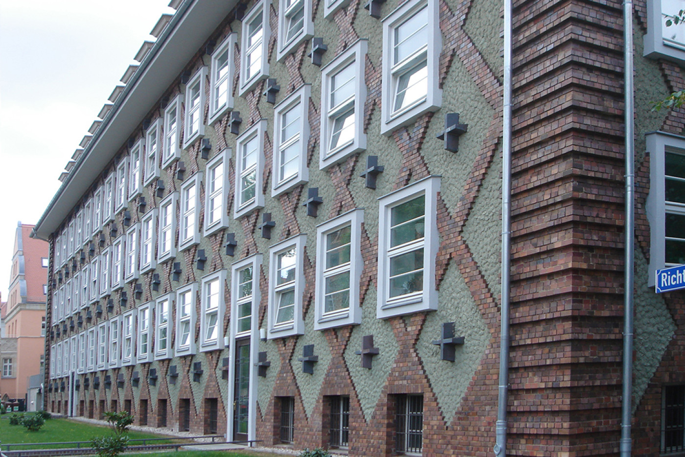 Schr&auml;gansicht der langen Fassade mit Fensterreihen, Rautenmuster aus Klinker und dunklen Konsolen, rechts die gestufte Klinkerkante.