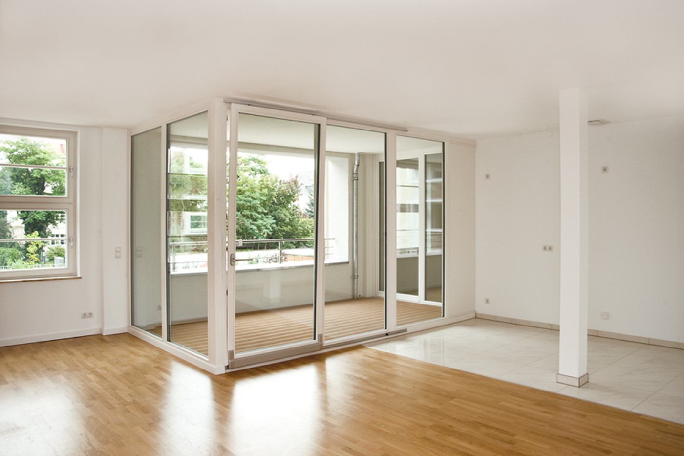 Leerer heller Wohnraum mit Parkettboden, verglaster Eckloggia und freistehender St&uuml;tze.