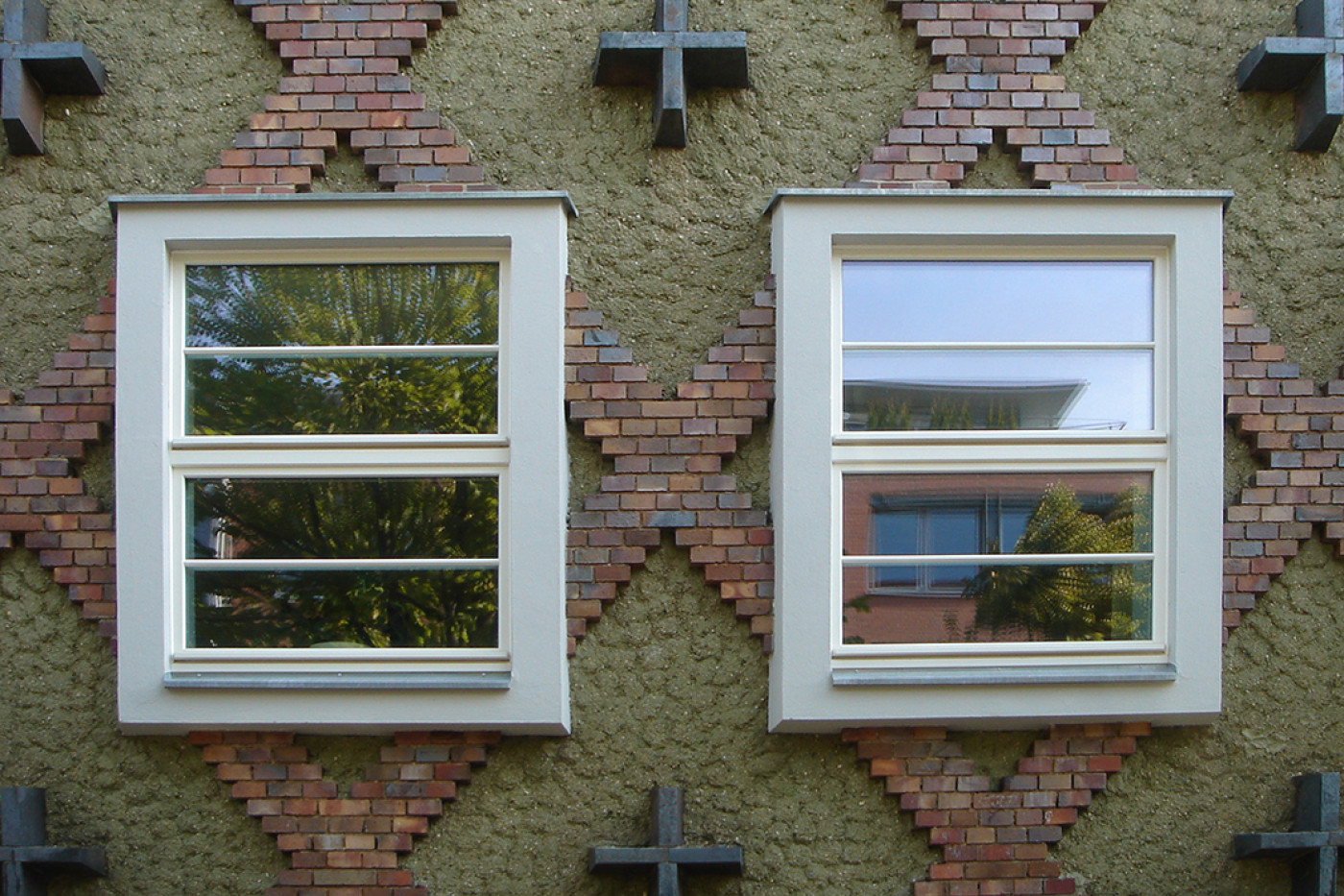 Zwei Sprossenfenster in der Rautenfassade mit kreuzf&ouml;rmigen Ankern, in den Scheiben spiegeln sich B&auml;ume.