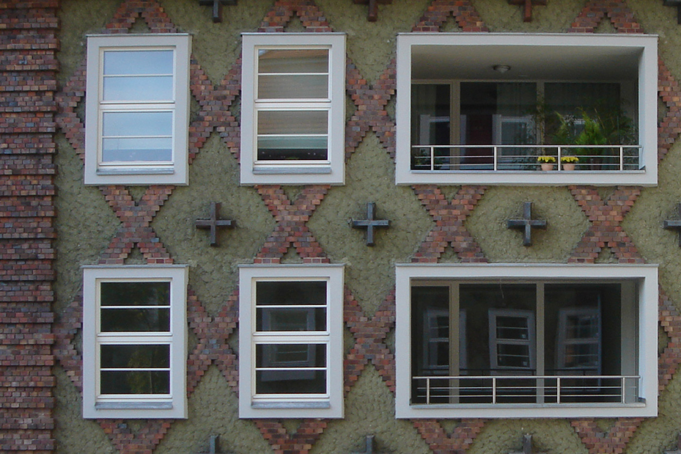 Fassadenausschnitt mit wei&szlig;en Fenstern und einer zur&uuml;ckgesetzten Loggia, dazwischen kreuzf&ouml;rmige Anker im Rautenmuster.