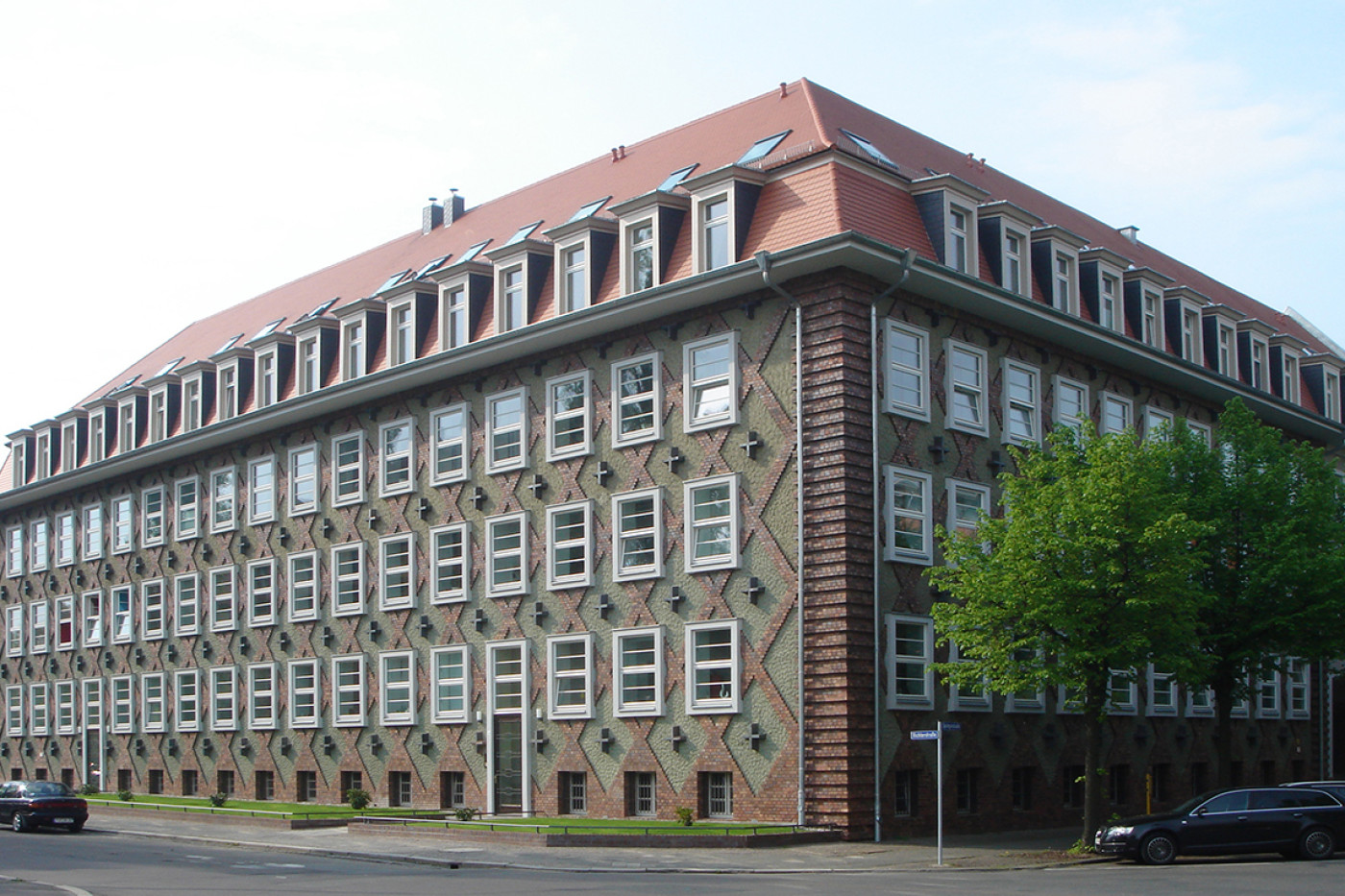 Eckansicht des f&uuml;nfgeschossigen Geb&auml;udes mit rotem Mansarddach und Dachgauben, die Fassade mit Rautenmuster aus Klinker und gr&uuml;nem Putz.