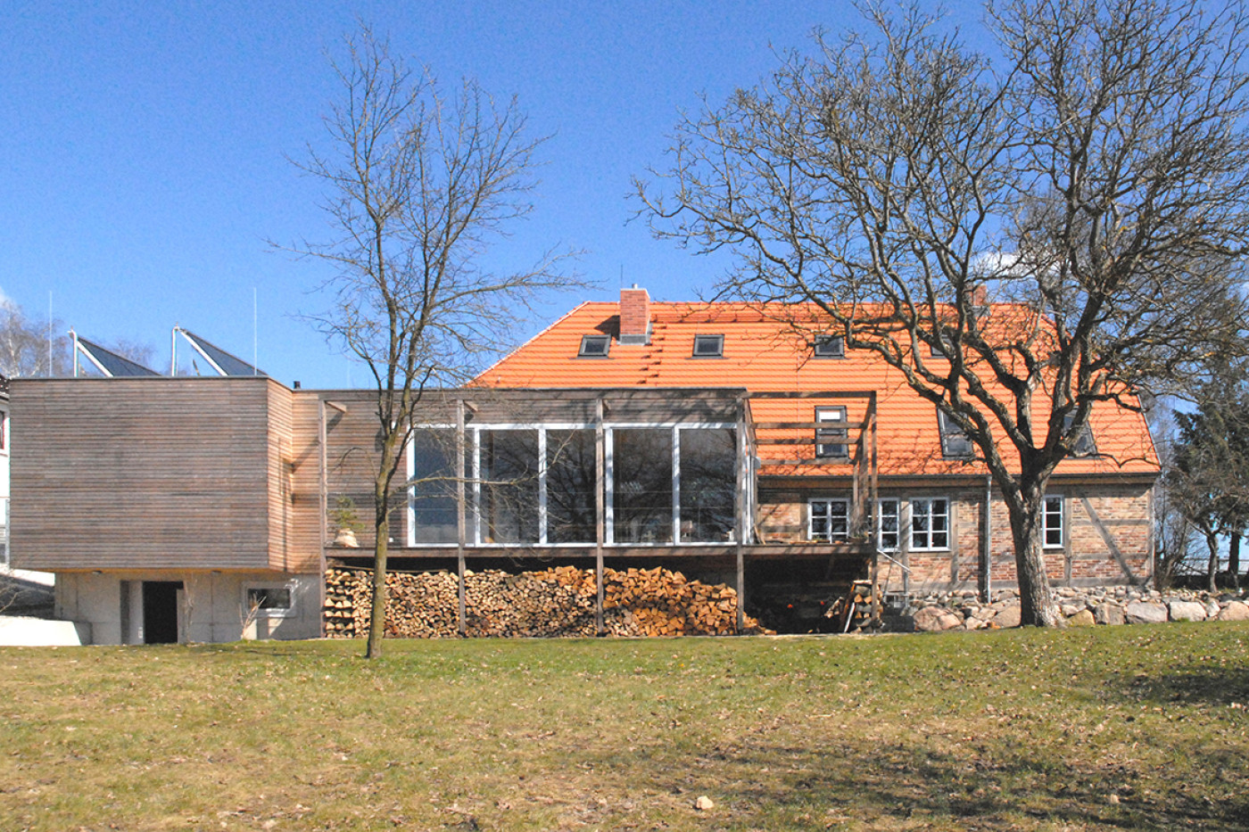 Das umgebaute Gasthaus von der Wiese aus: rotes Ziegeldach, verglaster Anbau und holzverkleideter Neubauteil links.