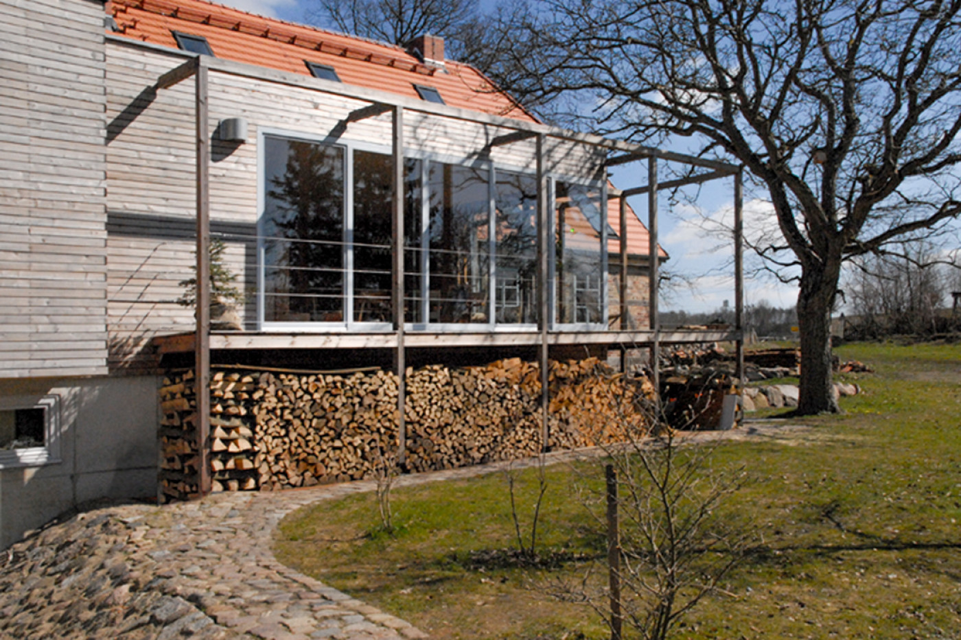 Verglaster Anbau mit h&ouml;lzerner Pergola und Terrasse, darunter gestapeltes Brennholz, davor ein gepflasterter Weg.