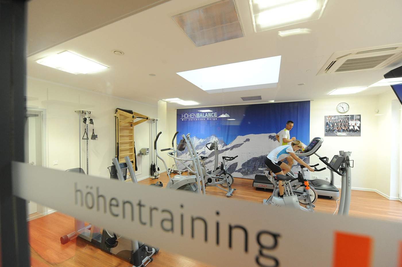 Blick durch eine beschriftete Glaswand mit dem Wort &bdquo;h&ouml;hentraining" auf Fahrradergometer vor einer Bergtapete.