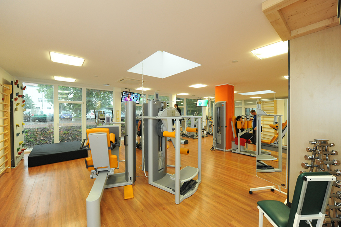 Trainingsraum mit orangefarbenen Ger&auml;ten, Dielenboden, Oberlicht und gro&szlig;en Fenstern zum Gr&uuml;n.