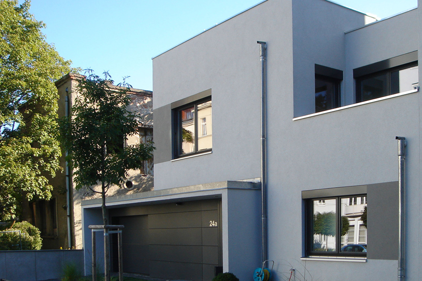 Grau verputzter Hausteil mit Garage, Hausnummer 24a und dunkel gerahmten Fenstern, davor ein junger Baum.