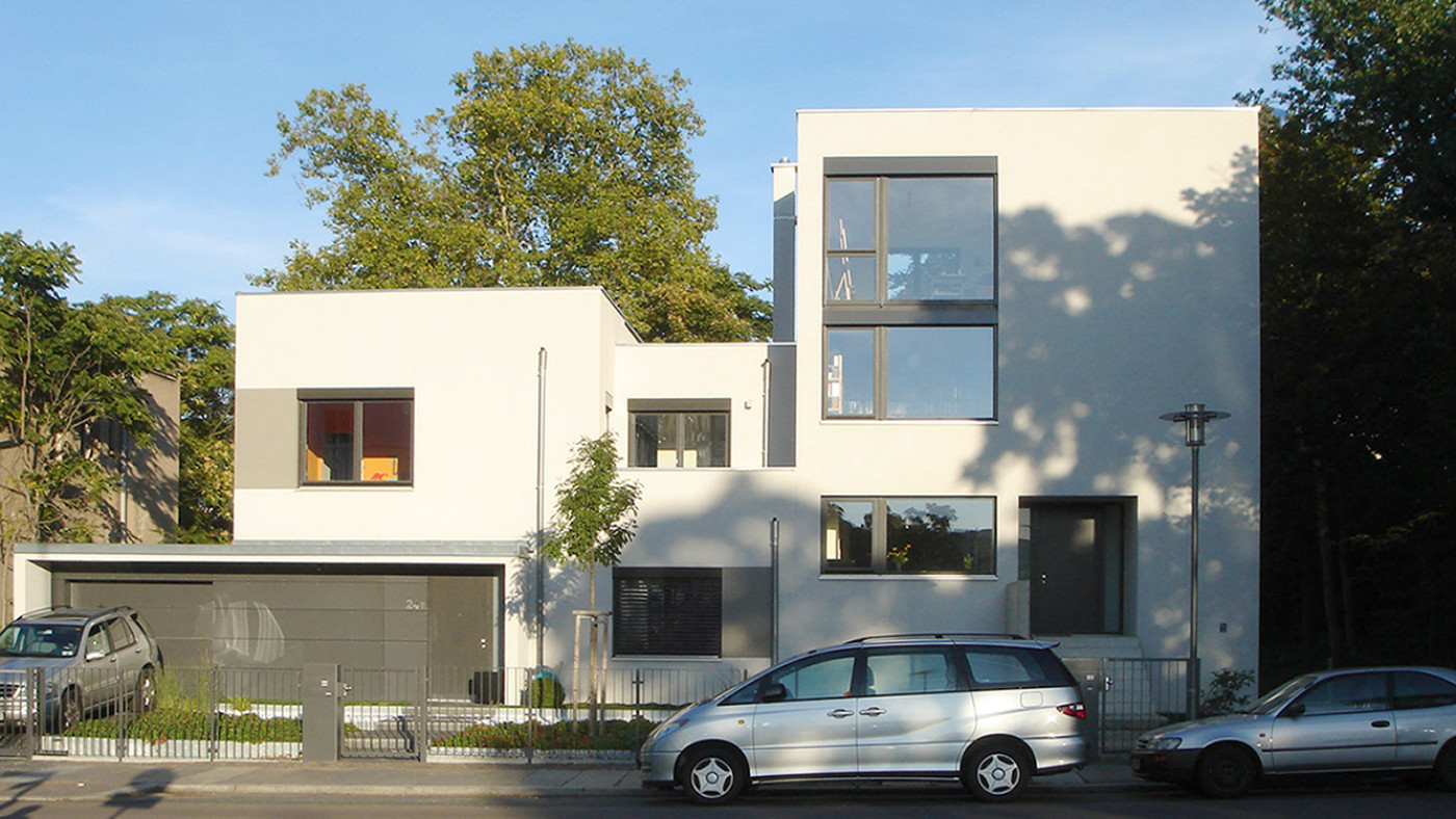 Wei&szlig;es Doppelhaus mit Flachdach und gro&szlig;en dunkel gerahmten Fenstern, links ein offener Carport.