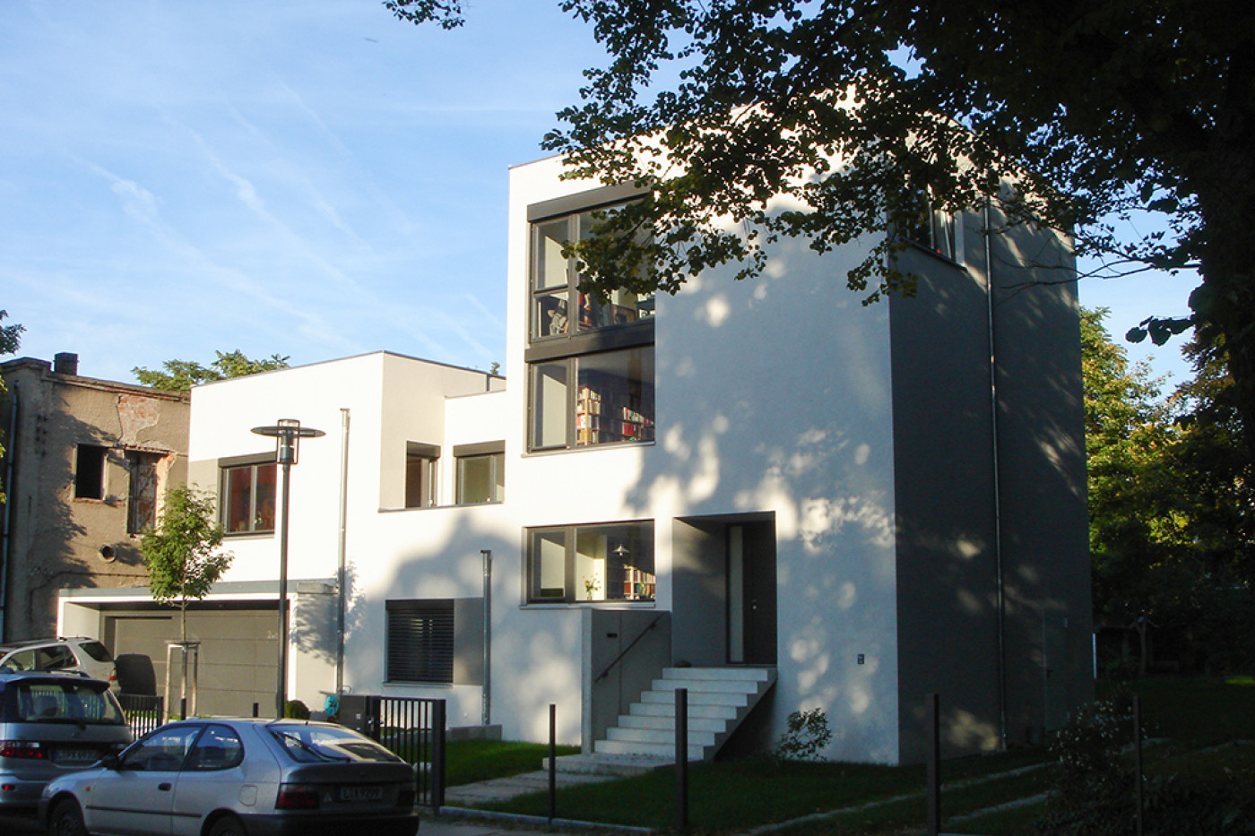 Wei&szlig;es Doppelhaus mit Flachdach und Freitreppe zum Eingang, dahinter B&auml;ume, davor die Stra&szlig;e.
