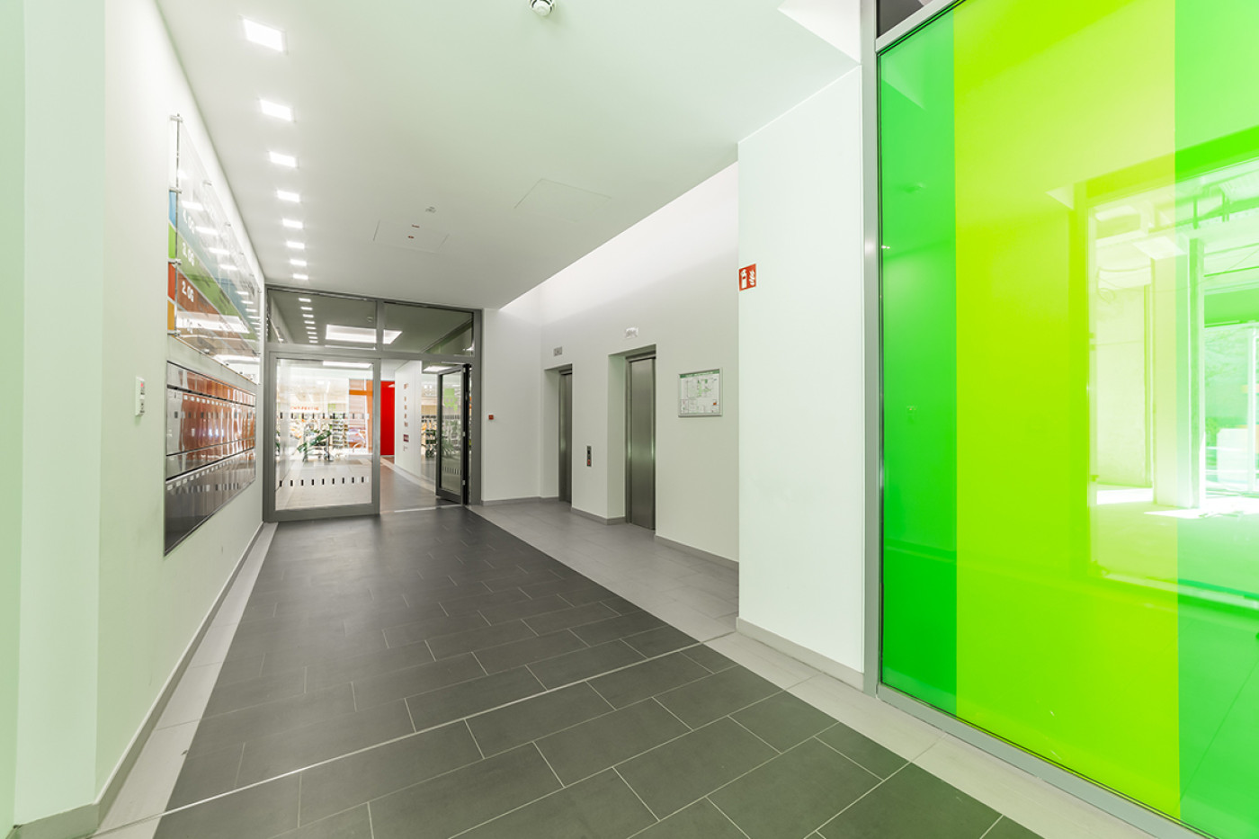 Flur mit leuchtend gr&uuml;ner Glaswand rechts und gerahmten Bildern an der linken Wand.