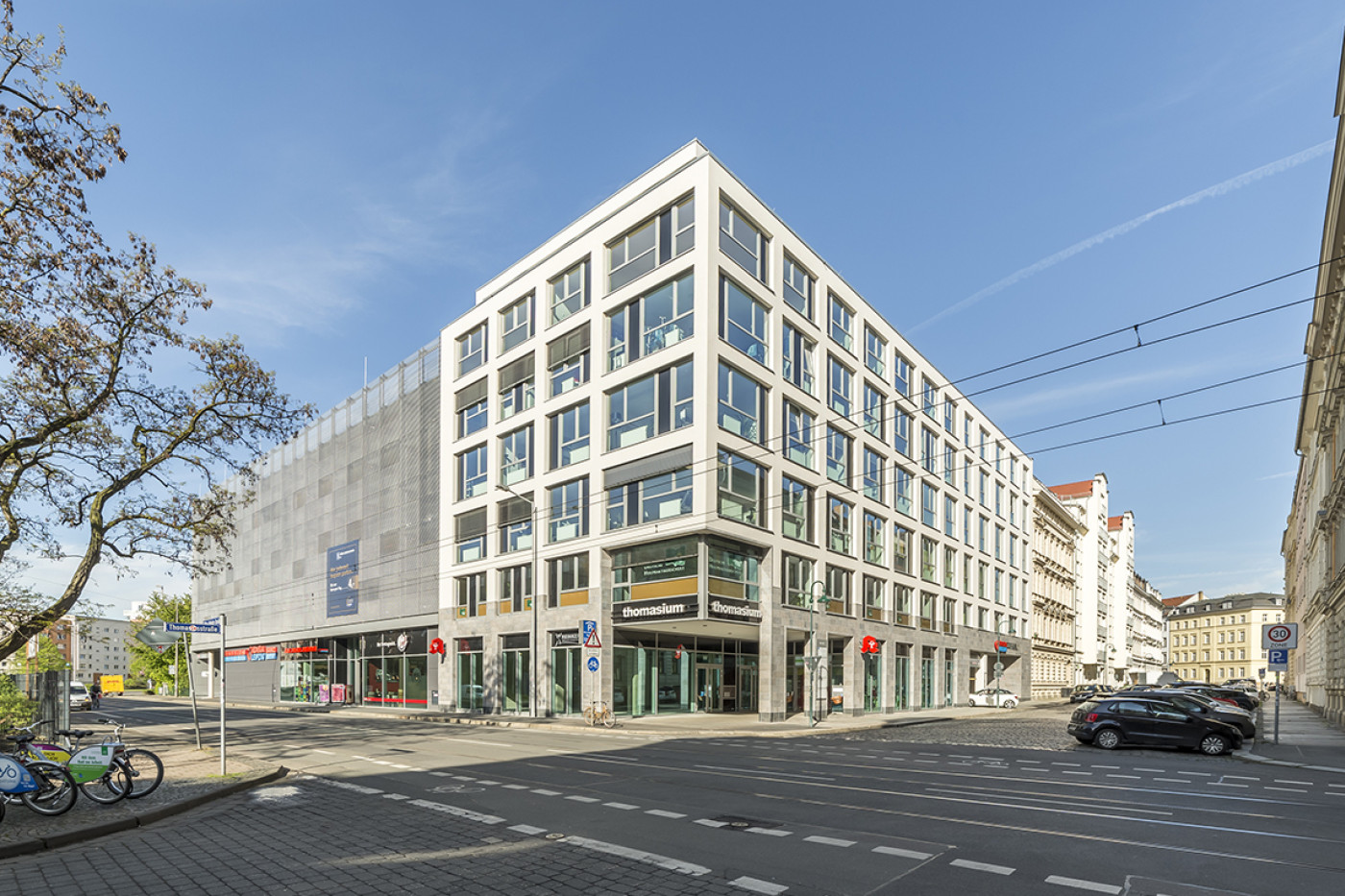 Eckgeb&auml;ude mit hellem Fassadenraster und verglastem Erdgeschoss an einer Kreuzung, links schlie&szlig;t das Parkhaus an.