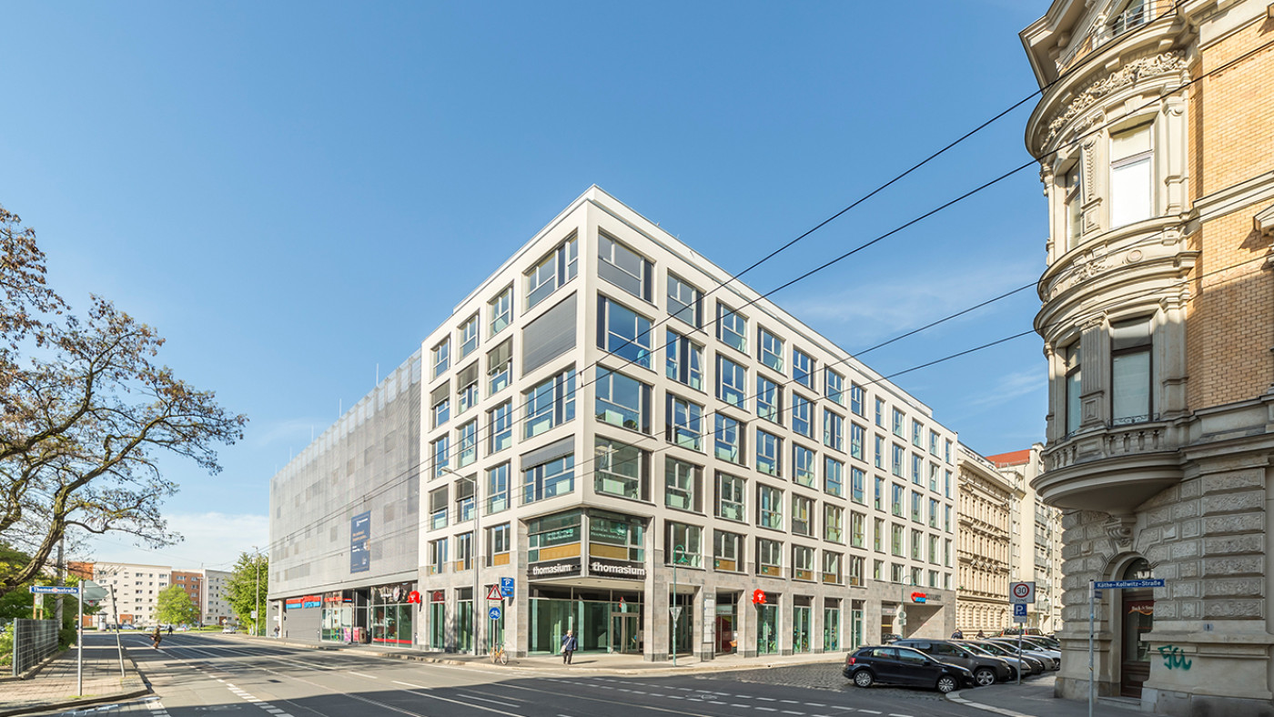 Eckgeb&auml;ude mit hellem Fassadenraster und Ladengeschossen, links schlie&szlig;t das Parkhaus an.