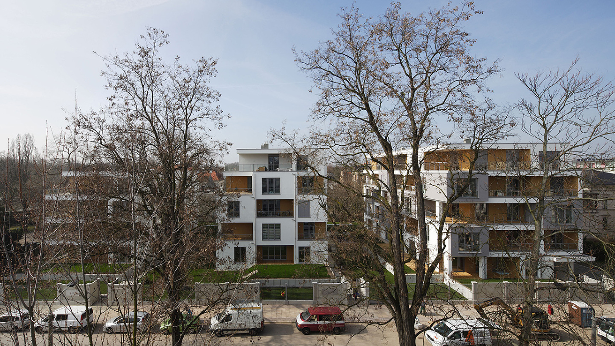Wei&szlig;e Wohnh&auml;user mit h&ouml;lzernen Balkonen, aufgenommen durch kahle B&auml;ume von der gegen&uuml;berliegenden Stra&szlig;enseite.