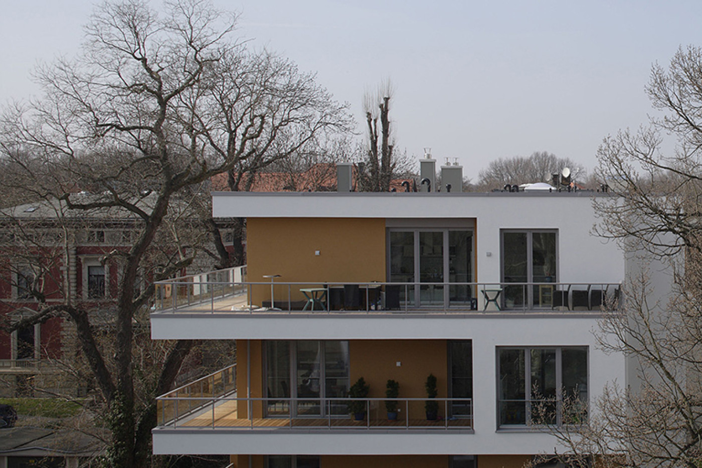 Kopfbau mit ockerfarben gefassten Balkonnischen und umlaufenden Br&uuml;stungen, links ein gro&szlig;er kahler Baum.