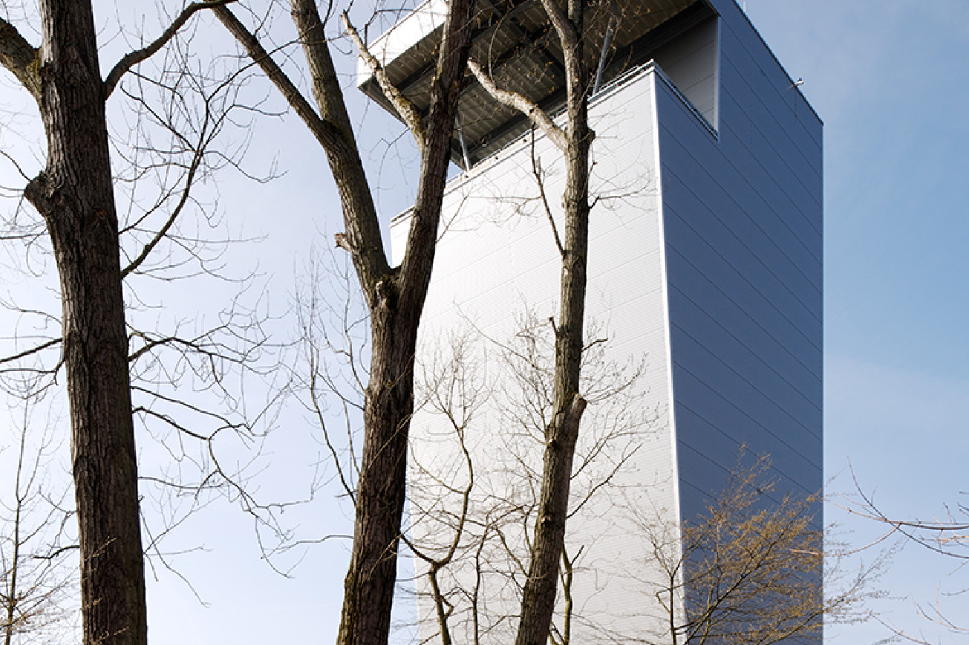 Schlanker &Uuml;bungsturm mit metallisch gl&auml;nzender Fassade und auskragender Plattform, davor kahle B&auml;ume.
