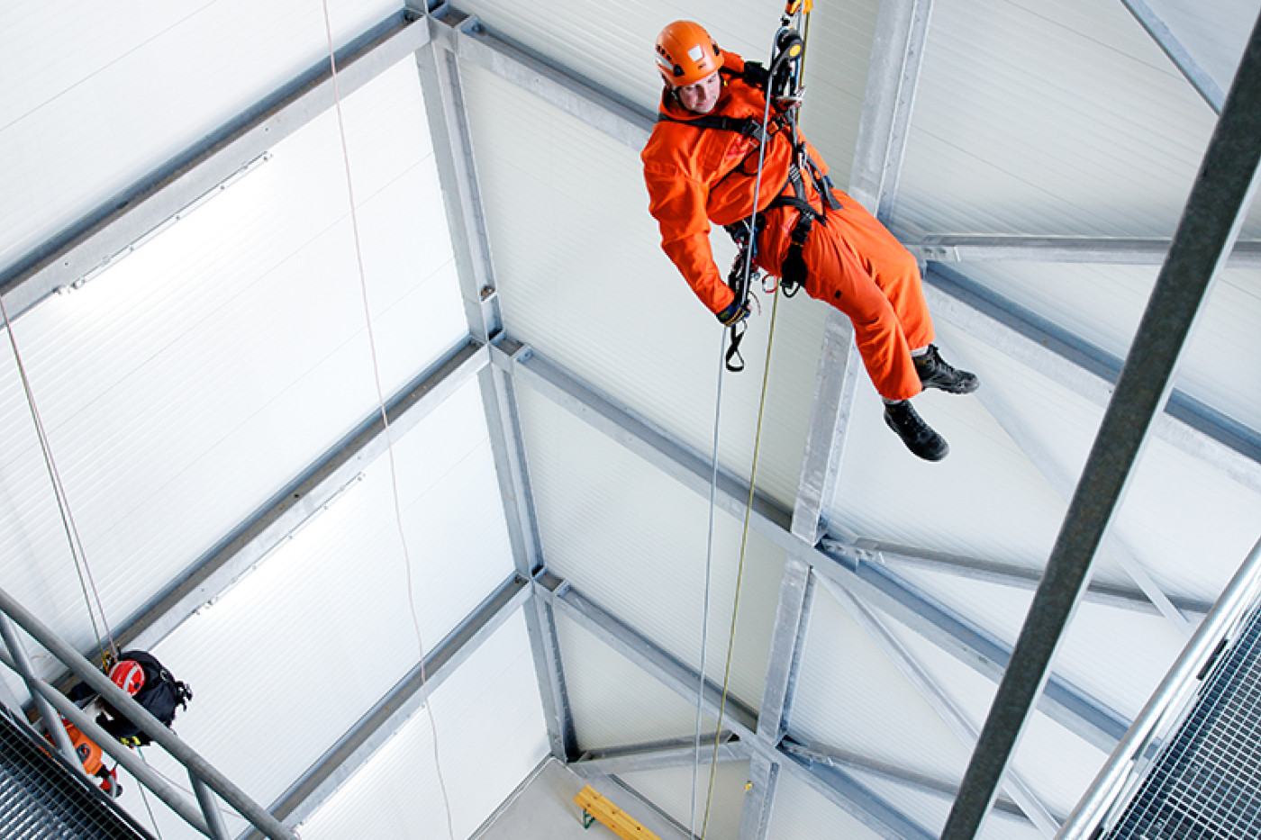 Blick von oben in die Halle des Turms: eine Person in orangefarbener Schutzkleidung h&auml;ngt frei im Seil.