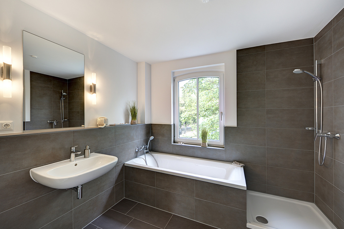 Bad mit graubraunen Fliesen, Waschbecken vor breitem Spiegel, Badewanne unter dem Fenster und bodengleicher Dusche.